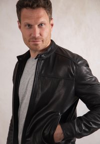 Black leather jacket with a zippered front, high collar, and minimal stitching details. Worn over a gray T-shirt, features angled side pockets.