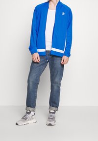 Sininen vetoketjullinen urheilutakki, jossa valkoiset yksityiskohdat, yllään valkoinen toppi, yhdistettynä käännettyihin farkkuihin ja harmaisiin lenkkareihin.