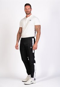 Weißes Poloshirt mit kurzem Kragen; schwarze Sporthosen mit weißen Seitenstreifen; weiße Sneakers; strukturierter Stoff; sichtbares Logo auf der Brust.