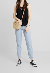 Un débardeur noir avec broderie florale, un jean bleu clair et des baskets en toile noires. Un sac bandoulière rond en paille avec une sangle marron est également présent.