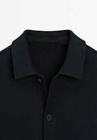 Camicetta nera con colletto, realizzata in un tessuto strutturato, con pattina dei bottoni e scollatura pulita, che presenta un design minimalista.