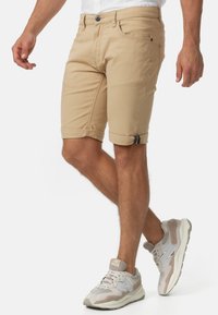 Beiga bomullsshorts med upprullade benslut, som har framfickor och en enda bakficka, kombinerade med ljusgrå och beigefärgade sneakers.