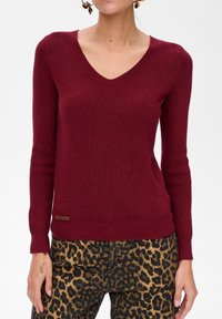 Bordowy sweter z prążkowanej dzianiny z dekoltem w serek, długimi rękawami i małą metką marki na dolnym brzegu. W połączeniu z spodniami w panterkę.