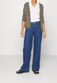 Zielony kardigan w paski nałożony na biały t-shirt w dekoltem w kształcie litery V, w połączeniu z niebieskimi spodniami typu wide-leg i czarnymi sandałami.