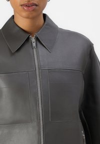 Graue Lederjacke mit kragenförmigem Ausschnitt, Frontreißverschluss und zwei großen Brusttaschen. Glatte Textur mit sauberen Nähten und minimalen Details.