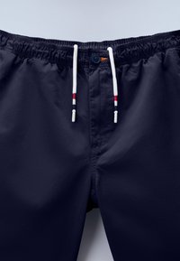 Μπλε σκούρα μαγιό σορτς με ελαστική μέση, άσπρα κορδόνια με κόκκινες και μαύρες ρίγες, και ένα μονο κουμπί κλεισίματος.