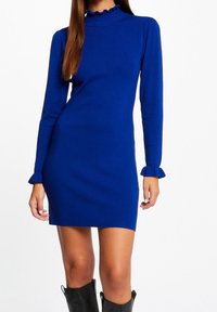 Mujer que lleva un vestido de punto entallado de manga larga en azul rey con puños y cuello con volantes, combinado con botas negras hasta la rodilla.