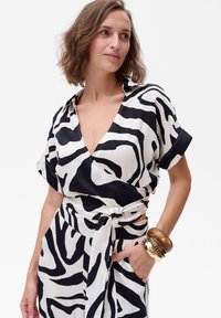 Zwart-wit gestreepte jurk met een V-hals en korte mouwen, voorzien van een strikceintuur. Model draagt forse metalen armbanden om één pols.