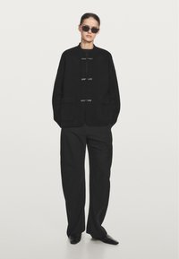 Giacca nera oversize con due tasche frontali, chiusura a bottoni e collo a giro. Abbinata a pantaloni neri a gamba larga e occhiali da sole rettangolari.