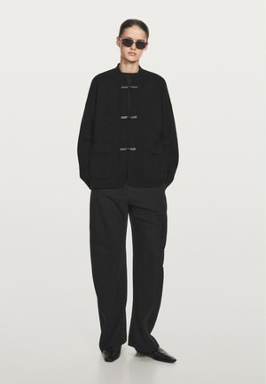 Zwarte oversized jas met twee voorkruimelpockets, knoopsluiting en ronde hals. Gecombineerd met wijde zwarte broeken en rechthoekige zonnebril.