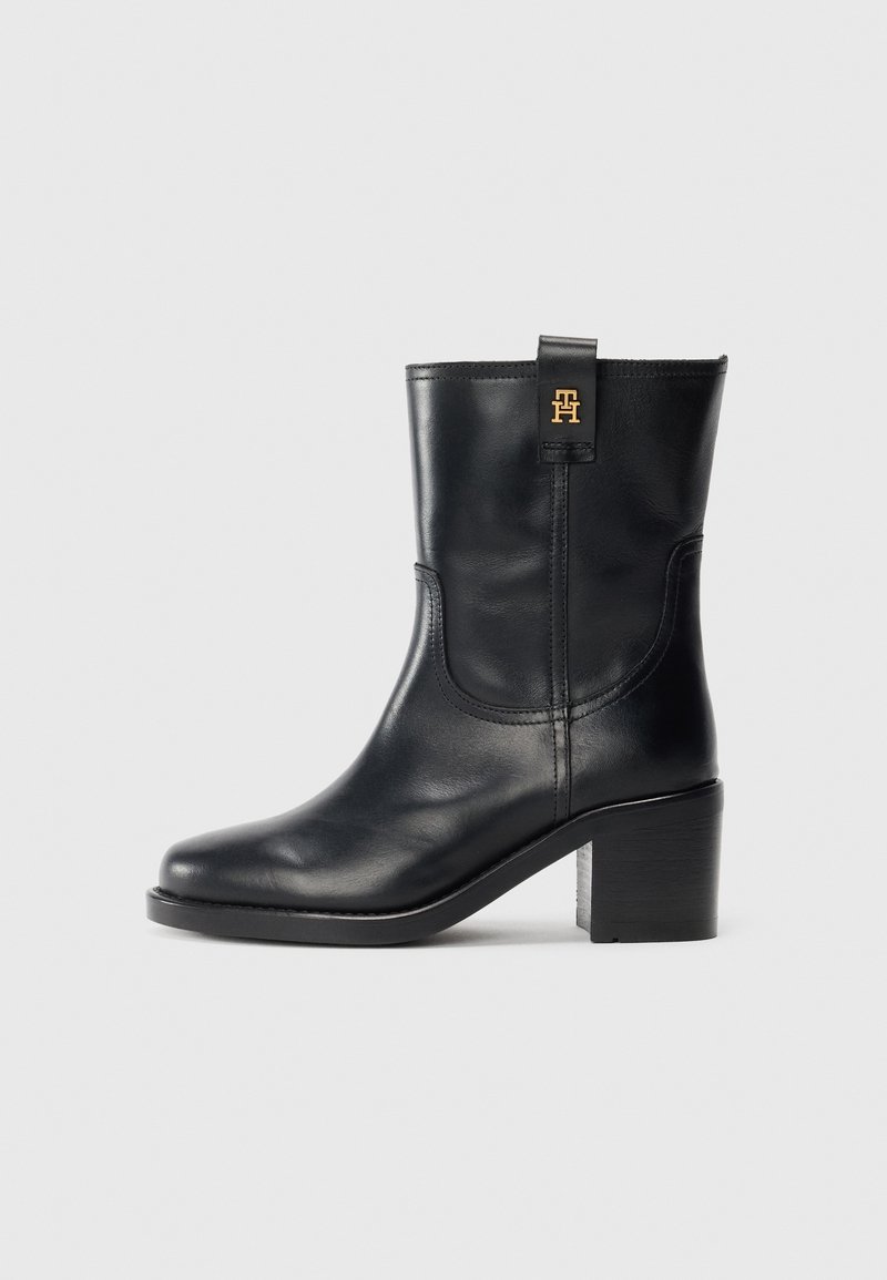 Bottine en cuir noir avec talon bloc, bout arrondi, languette latérale et petit logo doré sur le dessus, sur fond gris uni.