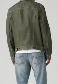 Grön denimjacka med ett strukturerat mönster, synliga sömnadsdetaljer och tryckknappar vid midjan. Bärs över ljusblå jeans.