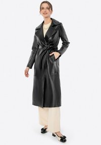 Cappotto trench in pelle nera con vita cinturata, colletto a revers e tasche laterali, abbinato a pantaloni crema e scarpe nere con fiocco.