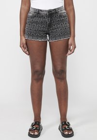 Melnas denim šortus ar izteiktu zīmējumu un saplēstu malu, kas ir atklāti ģeometriskā dizainā. Saskaņoti ar melnām sandalēm ar dekoratīvām sprādzēm.