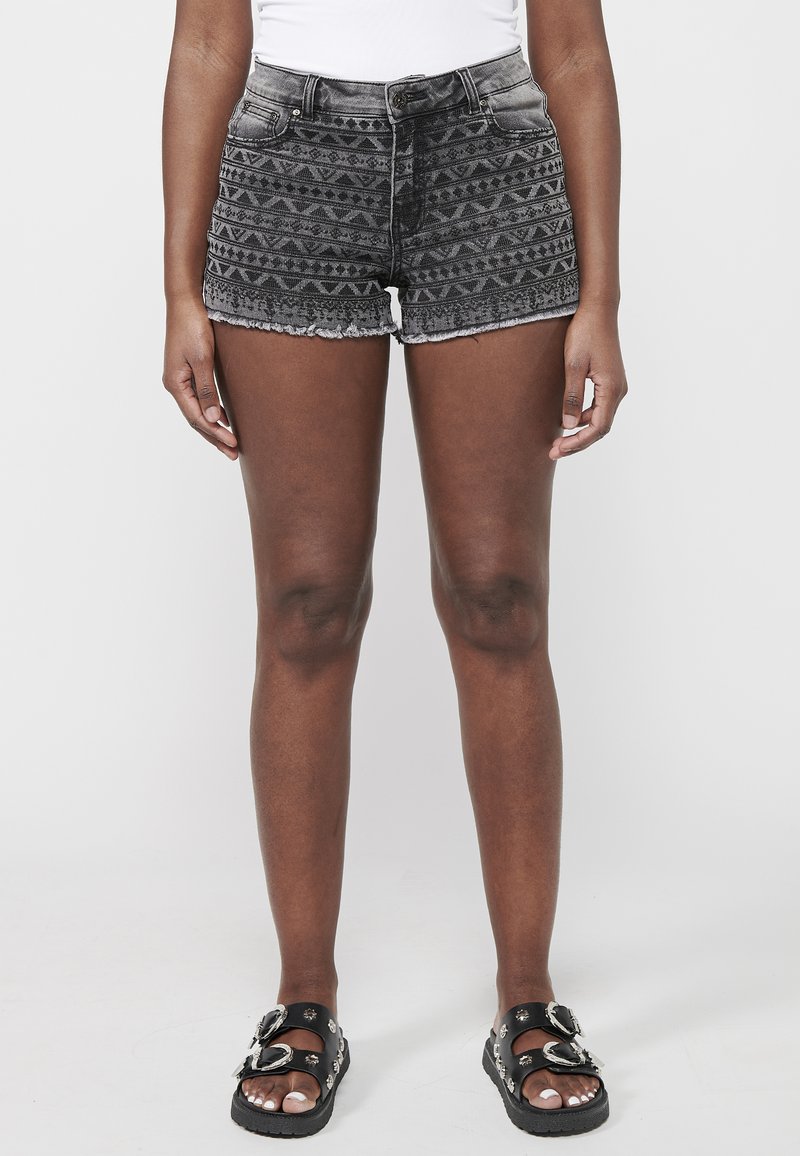 Melnas denim šortus ar izteiktu zīmējumu un saplēstu malu, kas ir atklāti ģeometriskā dizainā. Saskaņoti ar melnām sandalēm ar dekoratīvām sprādzēm.