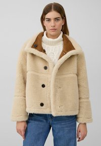 Beige schapenvachtjas met een bruine kraag, voorzien van vier zwarte knopen, wijde mouwen en een zachte textuur. Model draagt er een wit gebreide trui onder.