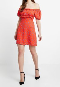 Šaty bez ramínek v jasně červené látce s malými modrými květinovými vzory, s upnutým pasem, krátkou sukní a volánkem na výstřihu.