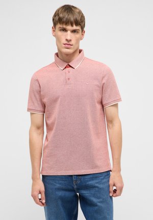 Junger Mann trägt ein hellrotes kurzärmeliges Polohemd mit weißem Besatz und blaue Jeans, steht vor einem schlichten weißen Hintergrund.