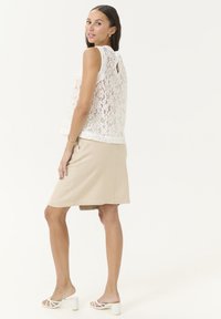 Haut en dentelle blanc avec une ouverture en forme de clé au dos, associé à une jupe beige arrivant au genou. Le modèle porte des sandales à talons compensés blanches. Texture lisse sur l'ensemble.