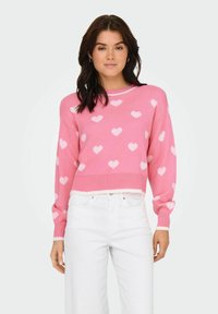 Roza skrajni pulover z belimi srčnimi vzorci, pletena tekstura, rebrasti manšeti in rob, v kombinaciji z belimi ravnimi hlačami.