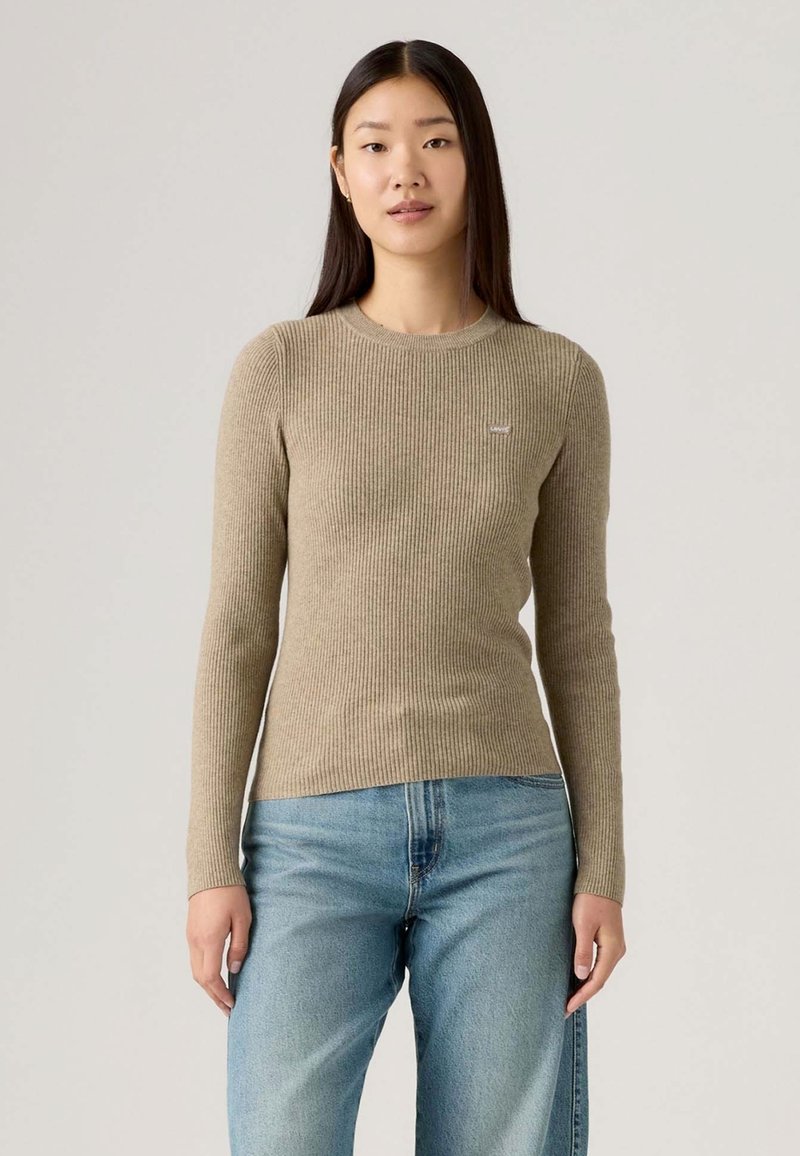 Frau mit langen dunklen Haaren, die einen beigefarbenen Rippenpullover und hellblaue Jeans trägt, steht vor einem schlichten hellgrauen Hintergrund.