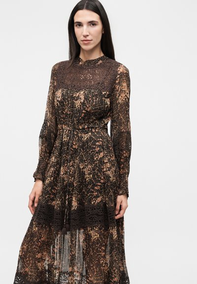 Γυναίκα με μακριά σκούρα μαλλιά φοράει διάφανο μαύρο και καφέ φόρεμα με λουλουδάτο σχέδιο και δαντέλα στο στήθος, τα μανίκια και το τελείωμα, στέκεται μπροστά σε απλό φόντο.