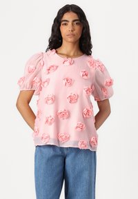 Blusa rosa a maniche corte con dettagli 3D a forma di rosa, scollatura rotonda e tessuto fluido, abbinata a jeans in denim blu chiaro.