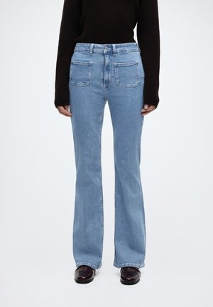 Άτομο που φοράει ανοιχτό μπλε τζιν ψηλόμεσο με εμπρόσθιες τσέπες, συνδυασμένο με μαύρο μακρυμάνικο τοπ και σκούρα παπούτσια, που στέκεται μπροστά σε λευκό φόντο.