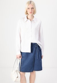 Hvit skjorte med broderte detaljer, kombinert med blå denim omslagskjørt. Modellen holder en hvit håndveske med buede hanker.