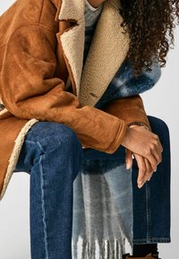 Brun, mockasinliknande kappa med en krämfärgad fårkrage, bärs över en blå mönstrad halsduk, tillsammans med blå denimjeans.