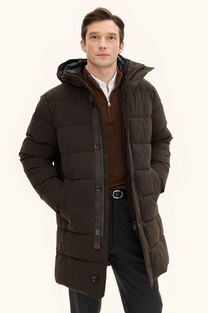 Langsamer brauner Steppjacke mit Kapuze, ausgestattet mit einem gesteppten Design, vorderen Knöpfen und einem Reißverschluss. getragen über einem braunen Pullover.