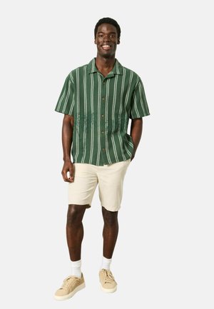 Lächelnder Mann, der ein grünes, gestreiftes Kurzarmhemd mit Knöpfen, beige Shorts, weiße Socken und beige Turnschuhe trägt und mit einer Hand in der Tasche steht.