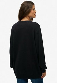 Czarny sweter z dzianiny o luźnym kroju, ściągacze przy mankietach i u dołu, teksturowana tkanina oraz okrągły dekolt. Widoczne detale ściegów na plecach.