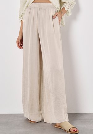 A woman wearing loose, light beige pants with an elastic waistband, paired with a cropped lace top and sandals on a gray surface.