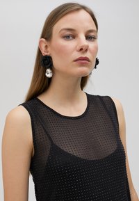 Svart ärmlös topp med en genomskinlig, prickig textur och rund halsringning; modellen bär svarta tygblommaörhängen och kristallaccent.