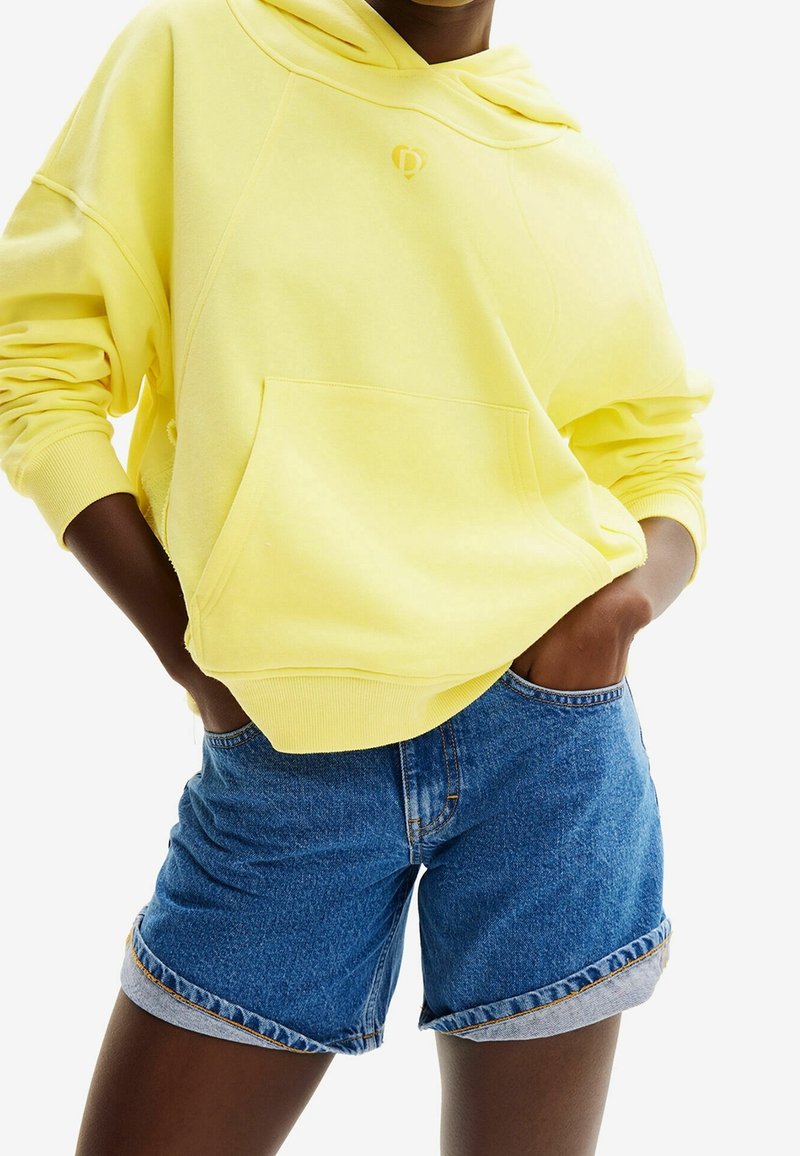 Personne portant un sweat à capuche jaune vif avec poche avant et short en jean bleu remonté, les mains dans les poches.