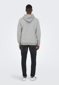 Felpa grigia in cotone con cappuccio regolabile, polsini e orlo a coste, abbinata a jeans scuri e sneakers bianche con dettagli verdi.