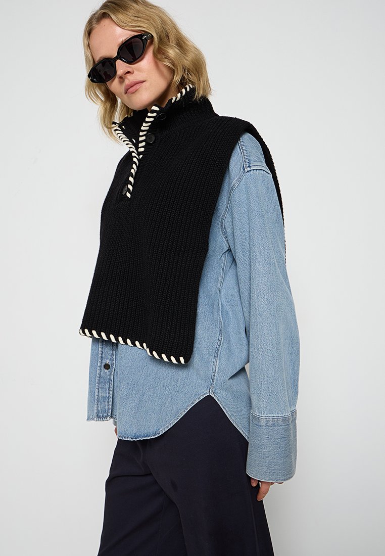 Schwarzes gestricktes ärmelloses Top mit hohem Kragen und weißen Kordelakzenten, über einem hellblauen Denim-Hemd mit großen Manschetten geschichtet.