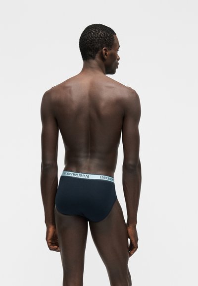 Άνδρας που στέκεται με την πλάτη στην κάμερα, φοράει μαύρα briefs Emporio Armani με λευκή ζώνη μέσης με το λογότυπο, σε απλό φόντο.