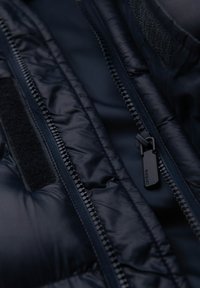 Vnitřek námořnické bundy s péřovou výplní ukazuje zip, suchý zip a lesklou tkaninu s prošívanou strukturou. Obsahuje plochý černý táhlo zipu.