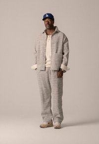 Casaco cinza claro texturizado com botões, combinado com calças do mesmo tecido. Camisa de malha bege por baixo e boné azul. Ténis castanhos completam o conjunto.