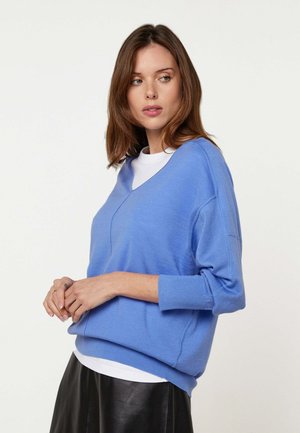 Femme aux cheveux bruns mi-longs portant un pull bleu ample à col en V sur un haut blanc et une jupe noire, regardant vers sa gauche.