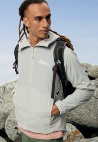 Lichtgrijze ritsjack met een capuchon, voorzien van voorzakken en een logo. Gelaagd over een lichtroze shirt, gedragen met een zwarte rugzak.