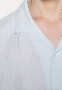 Světle modrá košile s krátkým rukávem z lnu, s jemnou texturou, špičatým límcem a předním zapínáním na knoflíky. Jemné variace v barvě.
