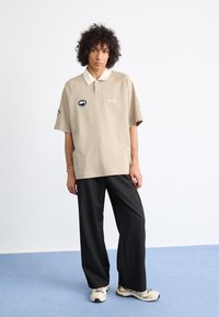 Persona joven con cabello rizado, vistiendo una camiseta polo beige con logotipos, pantalones anchos negros y zapatillas crema, de pie sobre un suelo azul contra una pared blanca.