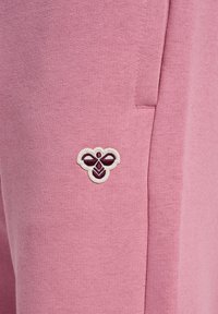 Pinkfarbener Stoff mit einem kleinen, dunkelroten und weißen bestickten Bienenlogo nahe einer vertikalen Naht, das die Textur und Nähdetails hervorhebt.