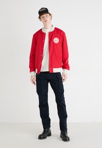 Red bomber jacket with white stripes at the cuffs and hem, featuring a circular patch. Paired with navy cargo pants and black boots.