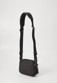 Černá crossbody taška s nastavitelným popruhem, elegantní obdélníkový design, hladká textura, zipové uzavření a kovové akcenty.
