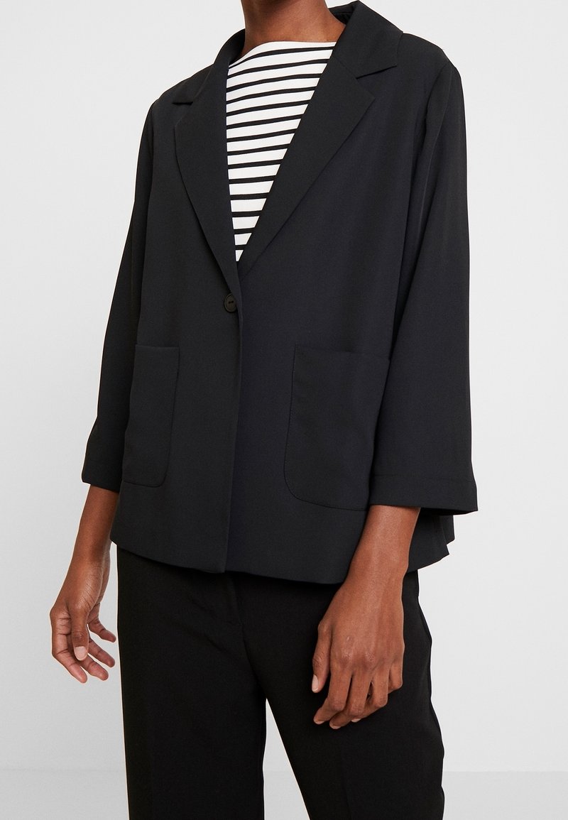 Blazer noir en tissu léger avec un col à revers cranté, une fermeture à un bouton unique et deux poches avant. Associé à un haut rayé en dessous.