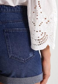 Valkoinen paita, jossa on monimutkaisella pitsisomisteella varustetut hihat, yhdistettynä sinisiin farkkushortseihin, joissa on klassinen takataskusuunnittelu ja k äännetty alaosa.